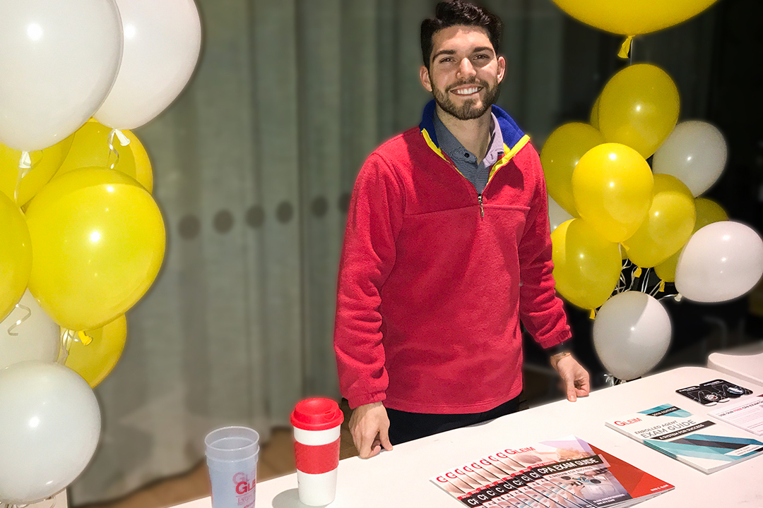 Austin at a tabling event, standing at a Gleim booth which is decorated with balloons and displays Exam Guides and Gleim merchandise.